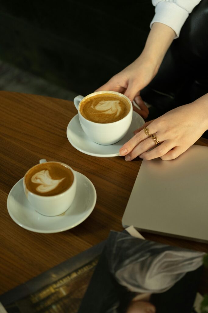 Hands hold coffee cups with latte art on a wooden table beside a laptop.